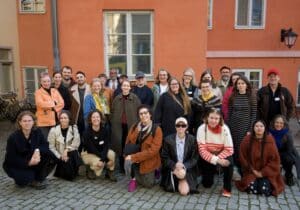 Group photo of about 30 people, men and women on the Turku Art House backyard, with an orange background.
